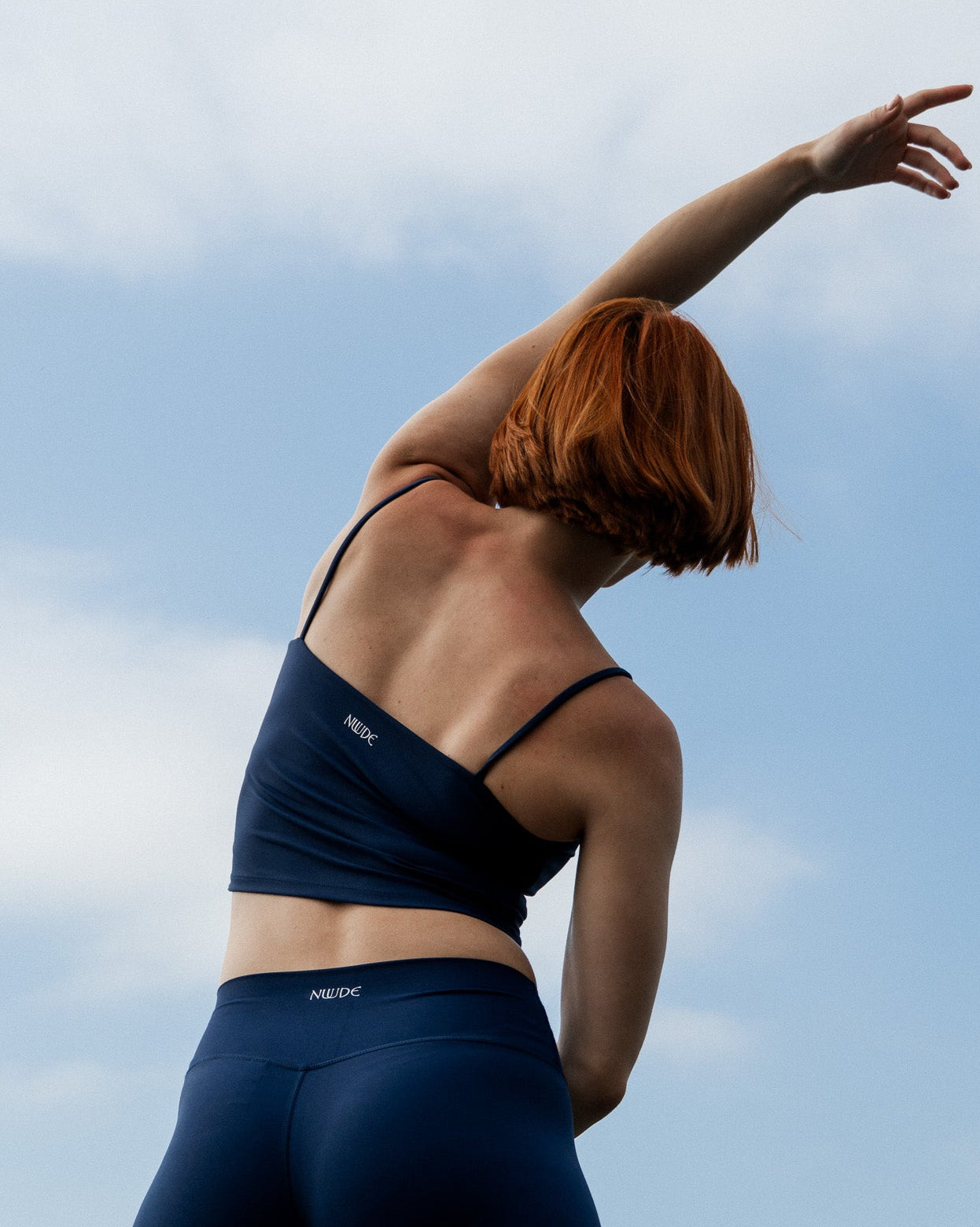 Person wearing a navy sports bra and leggings with 'Nuude' branding against a blue sky.