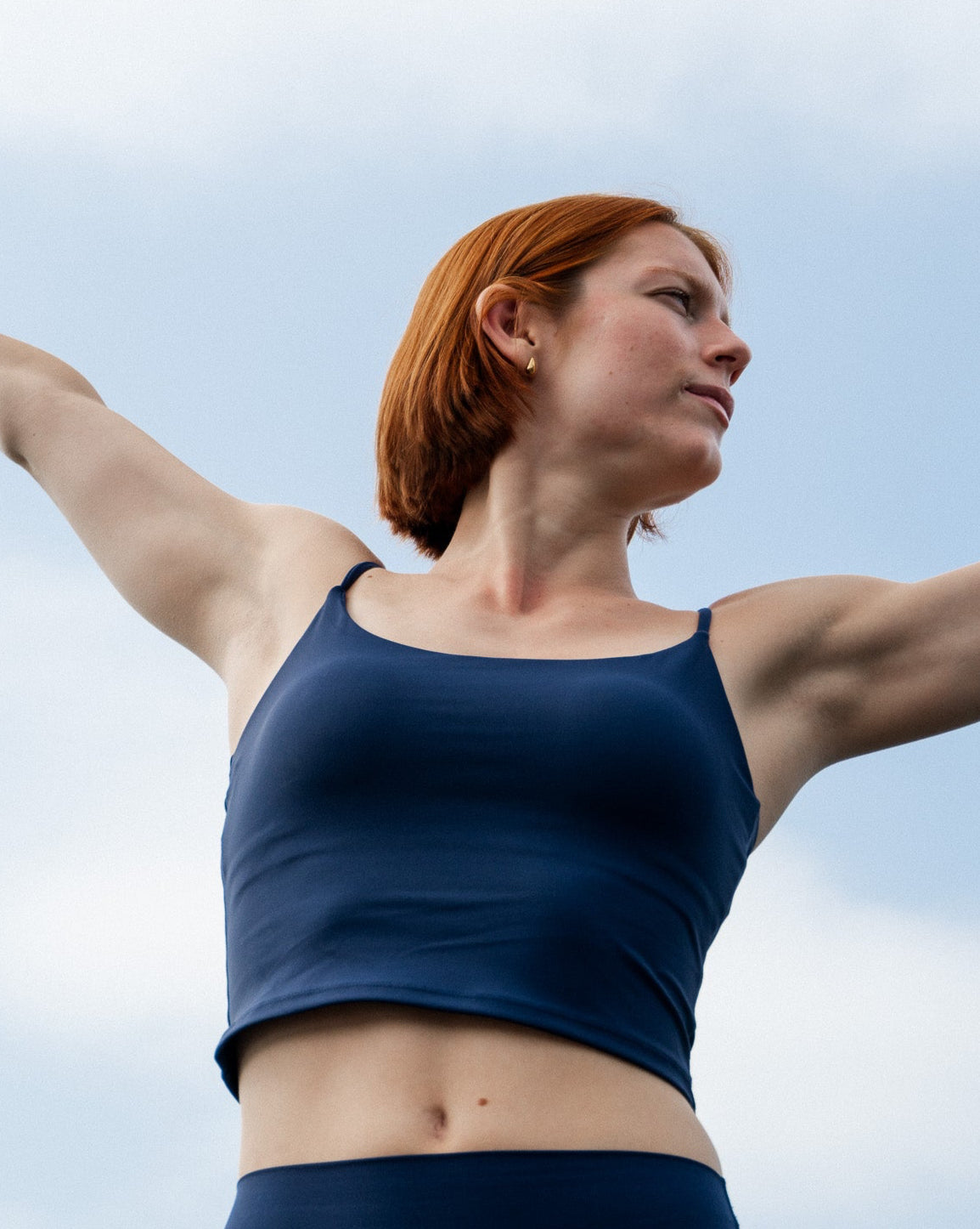 Woman in a blue sports bra and leggings with arms outstretched against a clear sky.